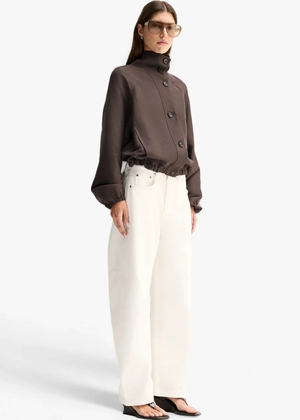 A model wearing a dark brown cropped button-up jacket with a high mock neck and gathered cuffs, paired with white trousers, against a light grey background.