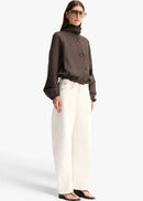 A model wearing a dark brown cropped button-up jacket with a high mock neck and gathered cuffs, paired with white trousers, against a light grey background.