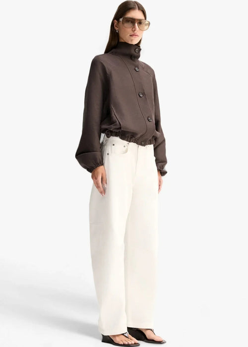 A model wearing a dark brown cropped button-up jacket with a high mock neck and gathered cuffs, paired with white trousers, against a light grey background.