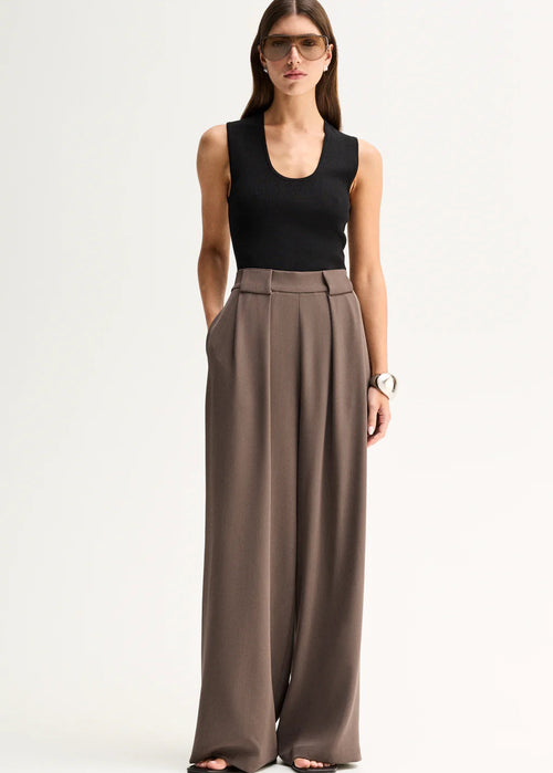 A woman modelling a sleeveless black top and high-waisted, wide-leg taupe-brown trousers against a plain white studio background.