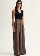A woman modelling a sleeveless black top and high-waisted, wide-leg taupe-brown trousers against a plain white studio background.