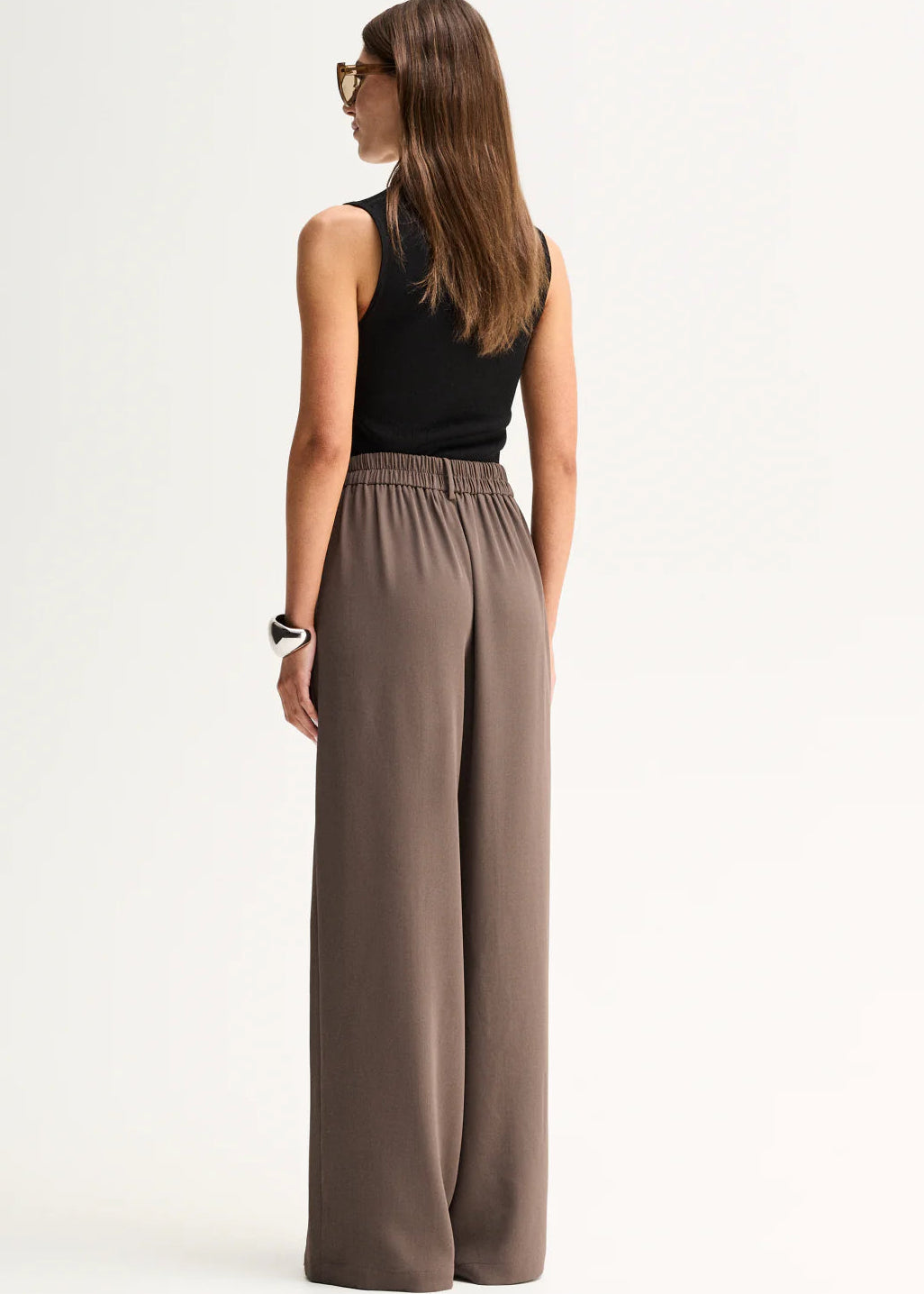 A woman modelling a sleeveless black top and high-waisted, wide-leg taupe-brown trousers against a plain white studio background.