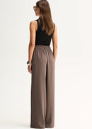 A woman modelling a sleeveless black top and high-waisted, wide-leg taupe-brown trousers against a plain white studio background.