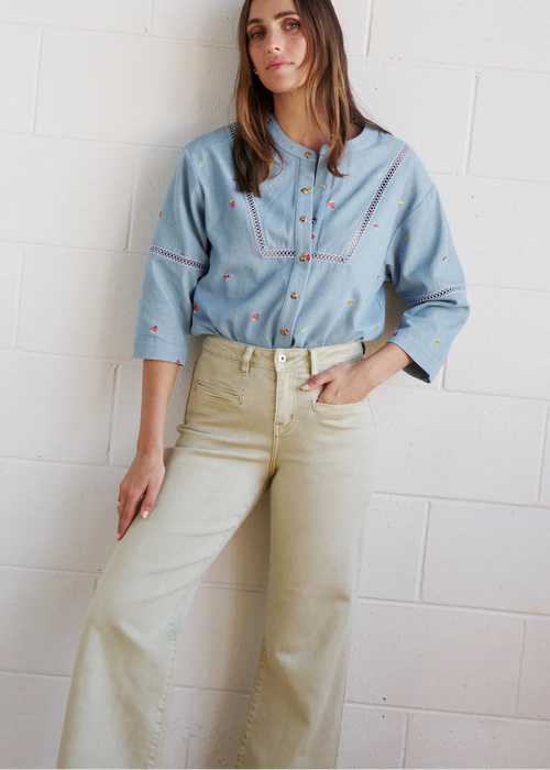 A woman wearing a light blue embroidered button-up shirt and beige wide-leg jeans, leaning against a white brick wall.