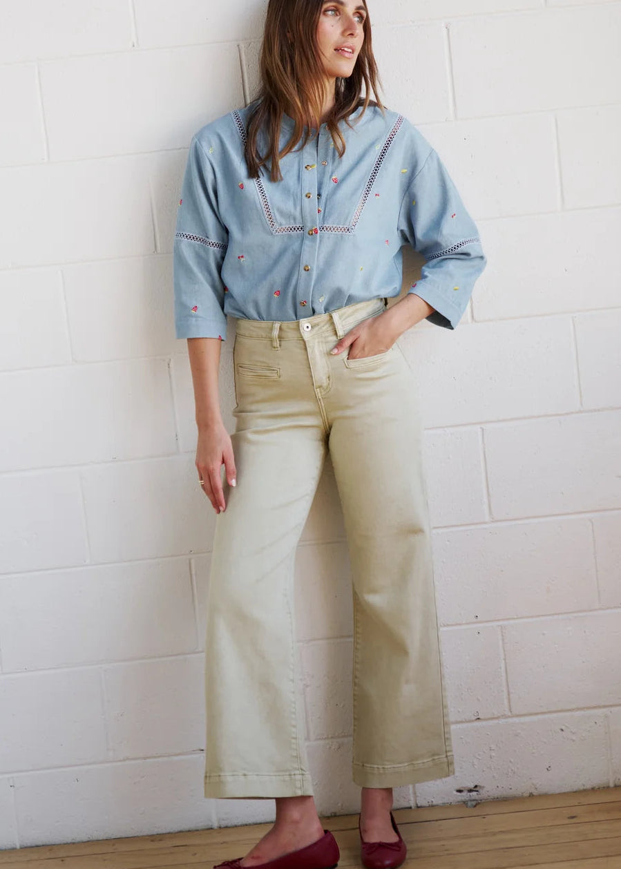 A woman wearing a light blue embroidered button-up shirt and beige wide-leg jeans, leaning against a white brick wall.