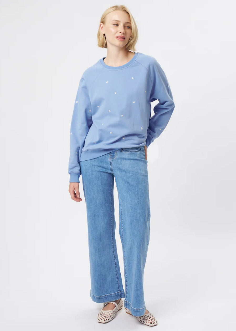 A woman modelling a light blue crewneck sweatshirt with small white embroidered details, paired with wide-leg light blue jeans and patterned flat shoes, against a plain white studio background.