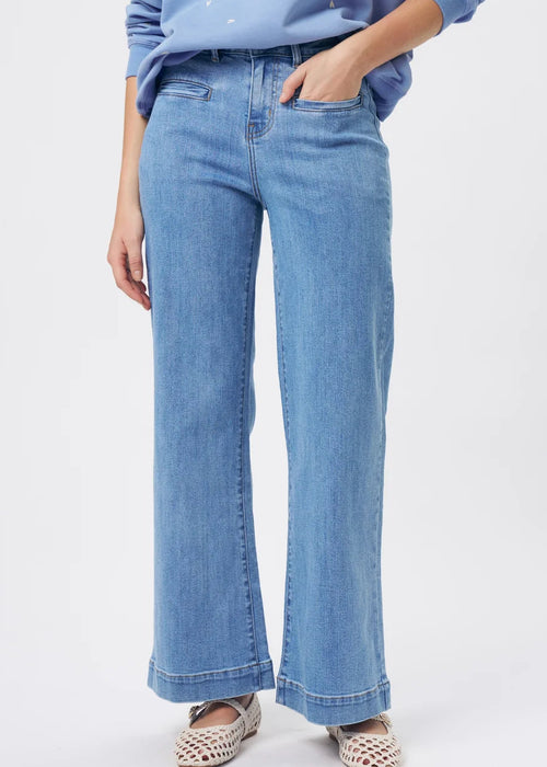 A woman modelling a light blue crewneck sweatshirt with small white embroidered details, paired with wide-leg light blue jeans and patterned flat shoes, against a plain white studio background.