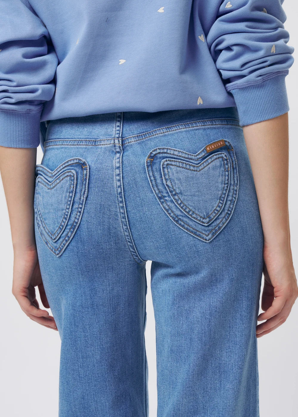 Close-up back view of a person wearing light blue jeans with heart-shaped back pockets and a light blue sweatshirt with small white embroidered details.
