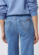 Close-up back view of a person wearing light blue jeans with heart-shaped back pockets and a light blue sweatshirt with small white embroidered details.