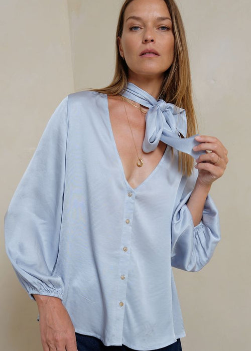 Woman wearing a light blue blouse with a scarf around her neck against a beige background