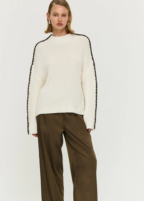 Woman wearing a white sweater with black trim and brown pants on a white background