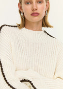 Woman wearing a white sweater with black trim and brown pants on a white background