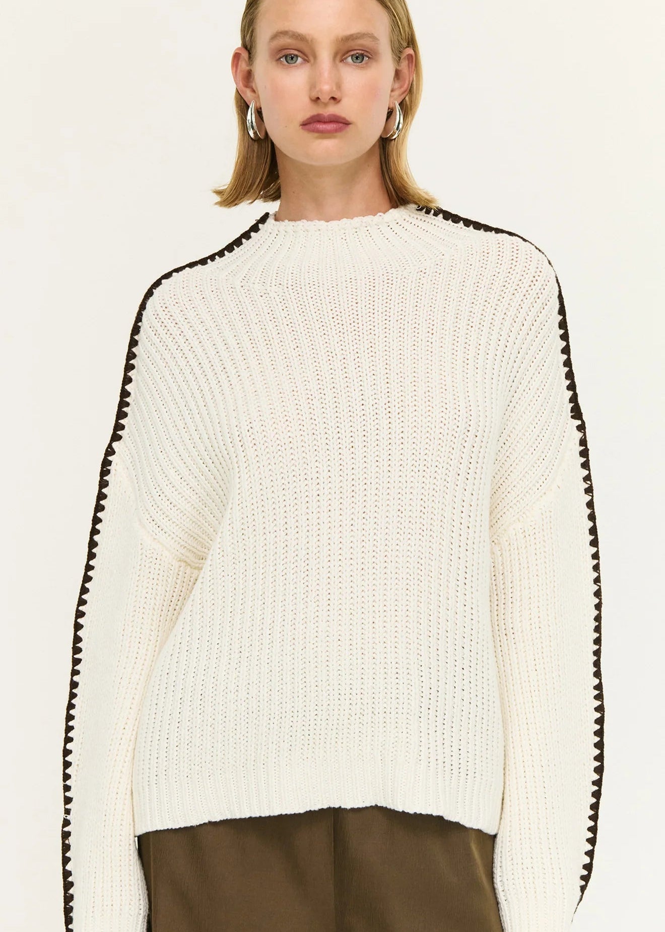 Woman wearing a white sweater with black trim and brown pants on a white background
