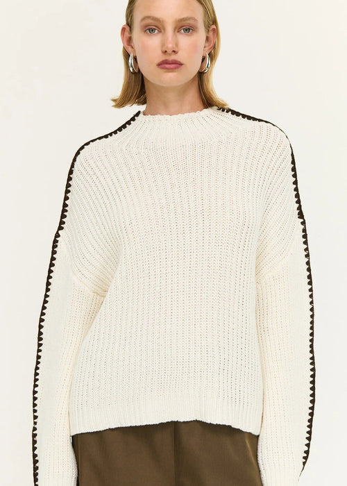 Woman wearing a white sweater with black trim and brown pants on a white background