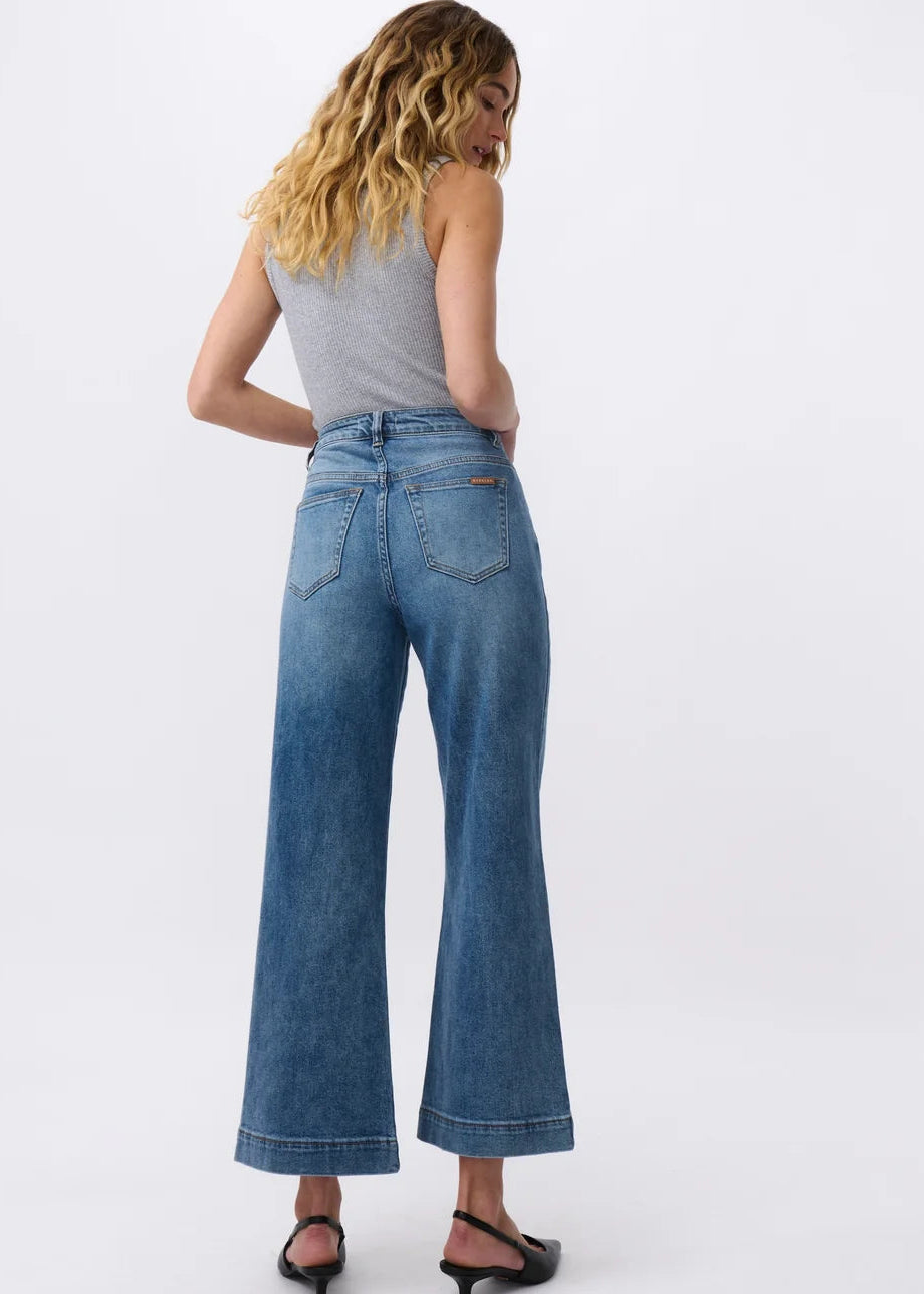 A woman shown from the back wearing high-waisted, wide-leg blue denim jeans, a grey sleeveless top, and black heeled sandals against a plain white studio background.