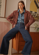Person wearing a red plaid shirt over a denim outfit, sitting on a wooden stool in a room with a poster on the wall.