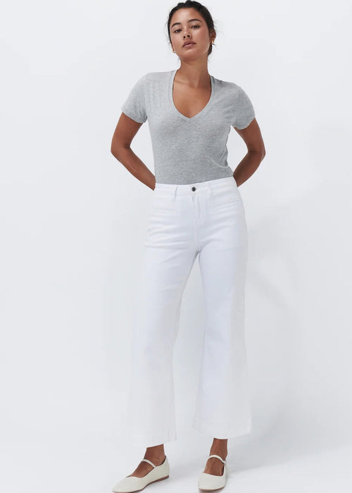 A woman modelling a light grey short-sleeve v-neck top and white cropped wide-leg jeans, paired with off-white Mary Jane flats, against a plain white studio background.