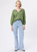 A woman modelling a sage green long-sleeve top with a split neckline and light-wash blue wide-leg jeans, standing against a plain white studio background.