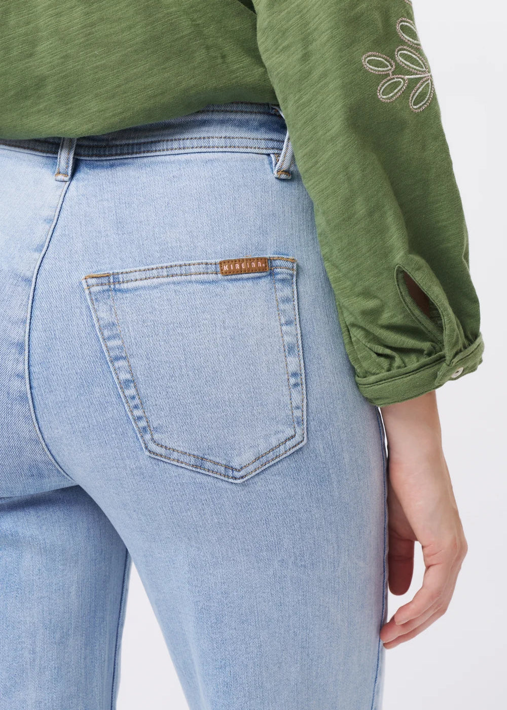 Close-up of a person wearing light-wash blue jeans and an olive green long-sleeve top, focusing on the back pocket area with a visible Fendi leather tab.