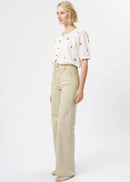 A blonde model is shown in profile wearing a white short-sleeve blouse with small red and green embroidered details, paired with high-waisted, wide-leg beige jeans and patterned shoes, against a plain white studio background.