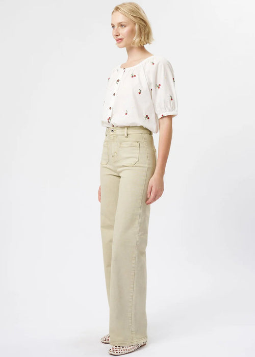 A blonde model is shown in profile wearing a white short-sleeve blouse with small red and green embroidered details, paired with high-waisted, wide-leg beige jeans and patterned shoes, against a plain white studio background.