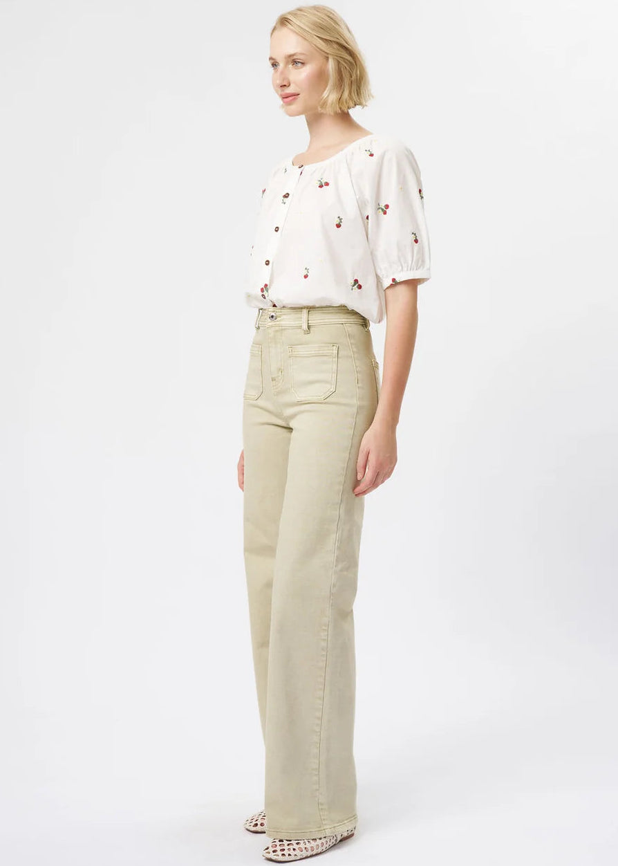 A blonde model is shown in profile wearing a white short-sleeve blouse with small red and green embroidered details, paired with high-waisted, wide-leg beige jeans and patterned shoes, against a plain white studio background.