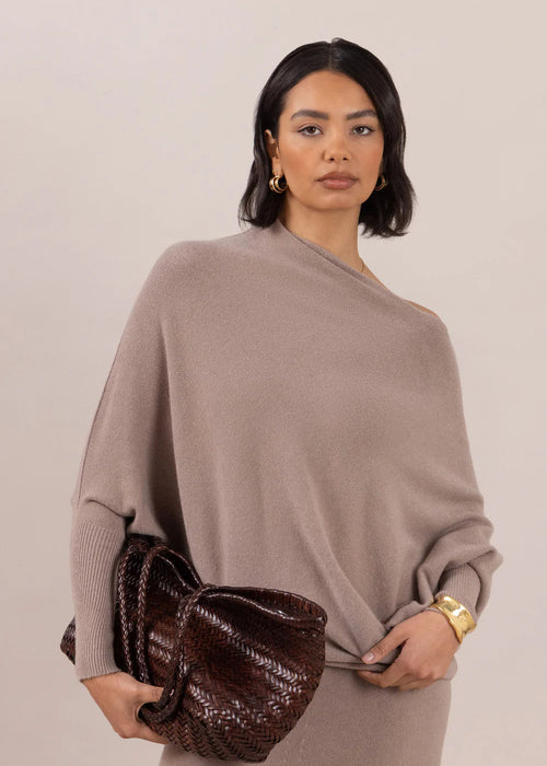 Woman wearing a beige sweater and pants holding a brown clutch against a plain background