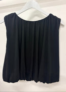 A black sleeveless top with a wide scoop neckline and gathered, draped fabric, hanging on a white hanger from a black clothing rack against a white wall.