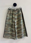 A pair of wide-leg shorts featuring a green and brown camouflage pattern with black side stripes, hanging from a black circular wall hook against a plain off-white wall.
