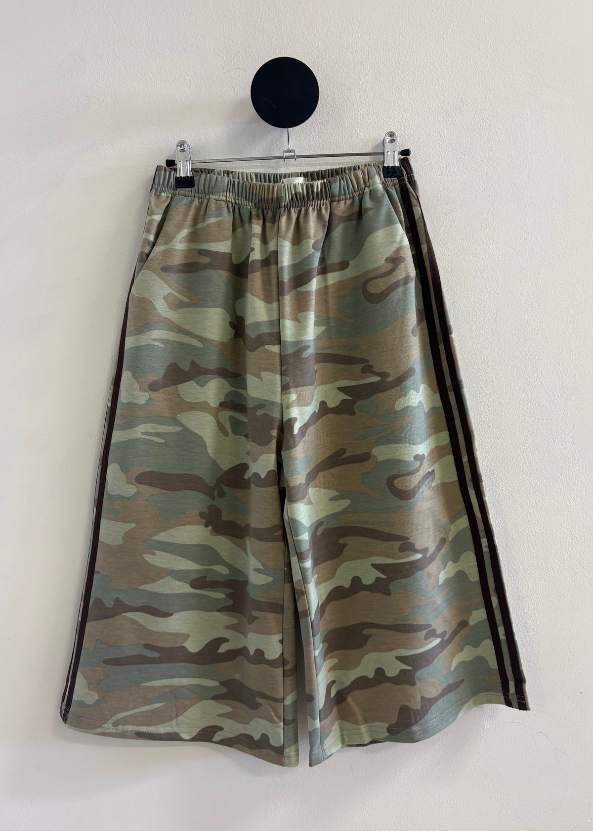 A pair of wide-leg shorts featuring a green and brown camouflage pattern with black side stripes, hanging from a black circular wall hook against a plain off-white wall.