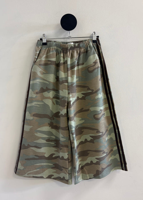 A pair of wide-leg shorts featuring a green and brown camouflage pattern with black side stripes, hanging from a black circular wall hook against a plain off-white wall.