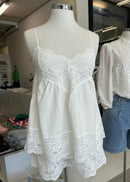 A white sleeveless camisole-style romper with lace trim displayed on a mannequin in a clothing store, alongside a matching white blouse and light blue denim shorts on another mannequin.