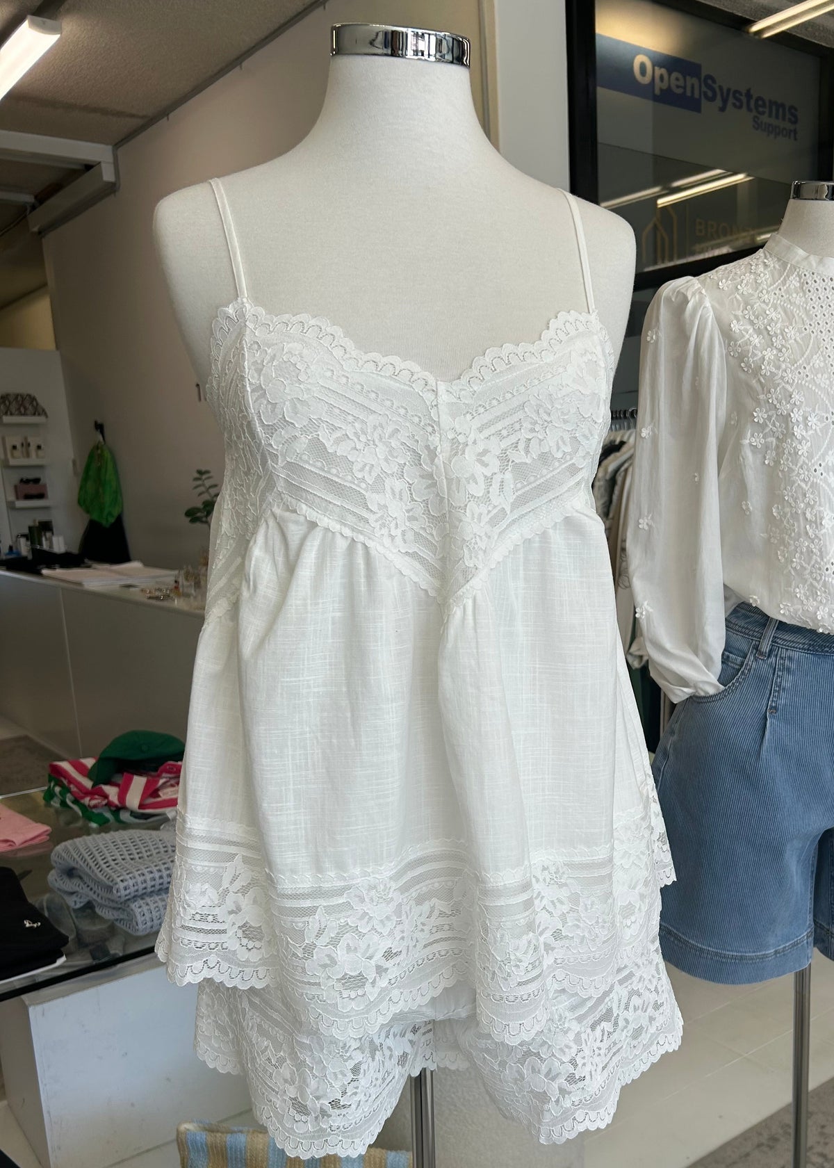 A white sleeveless camisole-style romper with lace trim displayed on a mannequin in a clothing store, alongside a matching white blouse and light blue denim shorts on another mannequin.