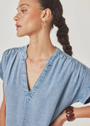 A woman wearing a light blue chambray short-sleeve blouse with a split V-neckline and gathered shoulders, shown in a close-up profile shot against a plain light background.
