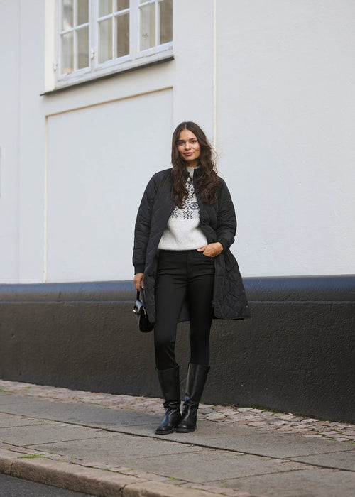 Woman in black coat and pants standing on a street with a white building in the background