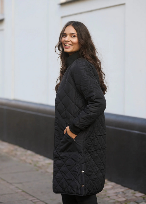 Woman in black coat and pants standing on a street with a white building in the background