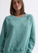 Woman wearing a teal sweater with small colourful embellishments on a white background