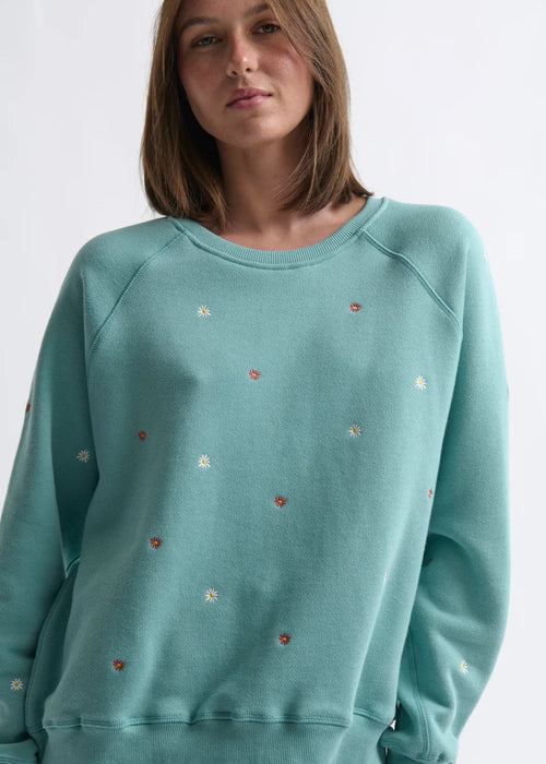 Woman wearing a teal sweater with small colourful embellishments on a white background