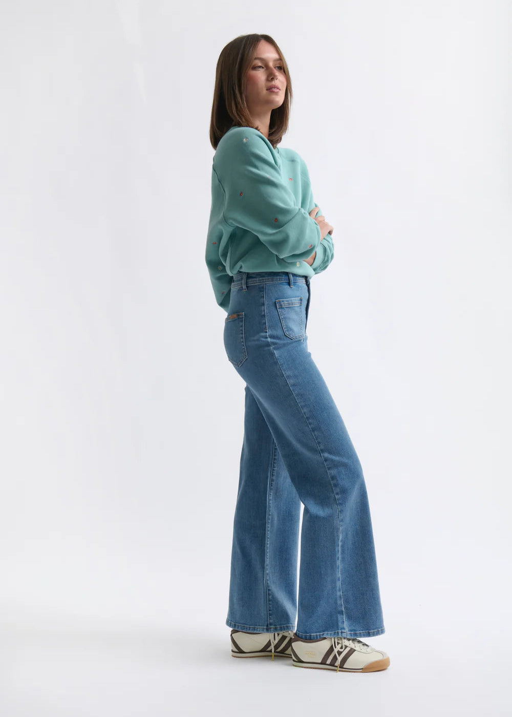 Woman wearing a teal sweater and blue jeans standing against a white background