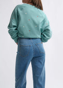 Person wearing a teal sweatshirt with small colourful patterns and blue jeans on a white background