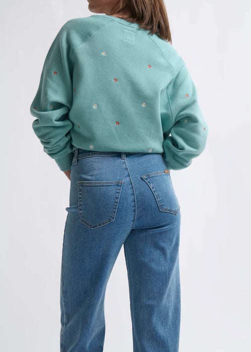 Person wearing a teal sweatshirt with small colourful patterns and blue jeans on a white background