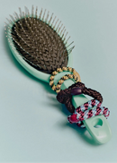 A mint green paddle hairbrush with dark brown bristles, decorated with three braided rope handles in gold, dark brown, and red-and-white patterned cords, set against a solid pale blue background.