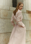 Woman in a long light pink coat standing against a stone wall.