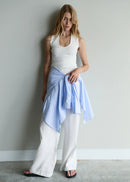 Woman wearing a white tank top and light blue scarf with white pants against a plain wall.