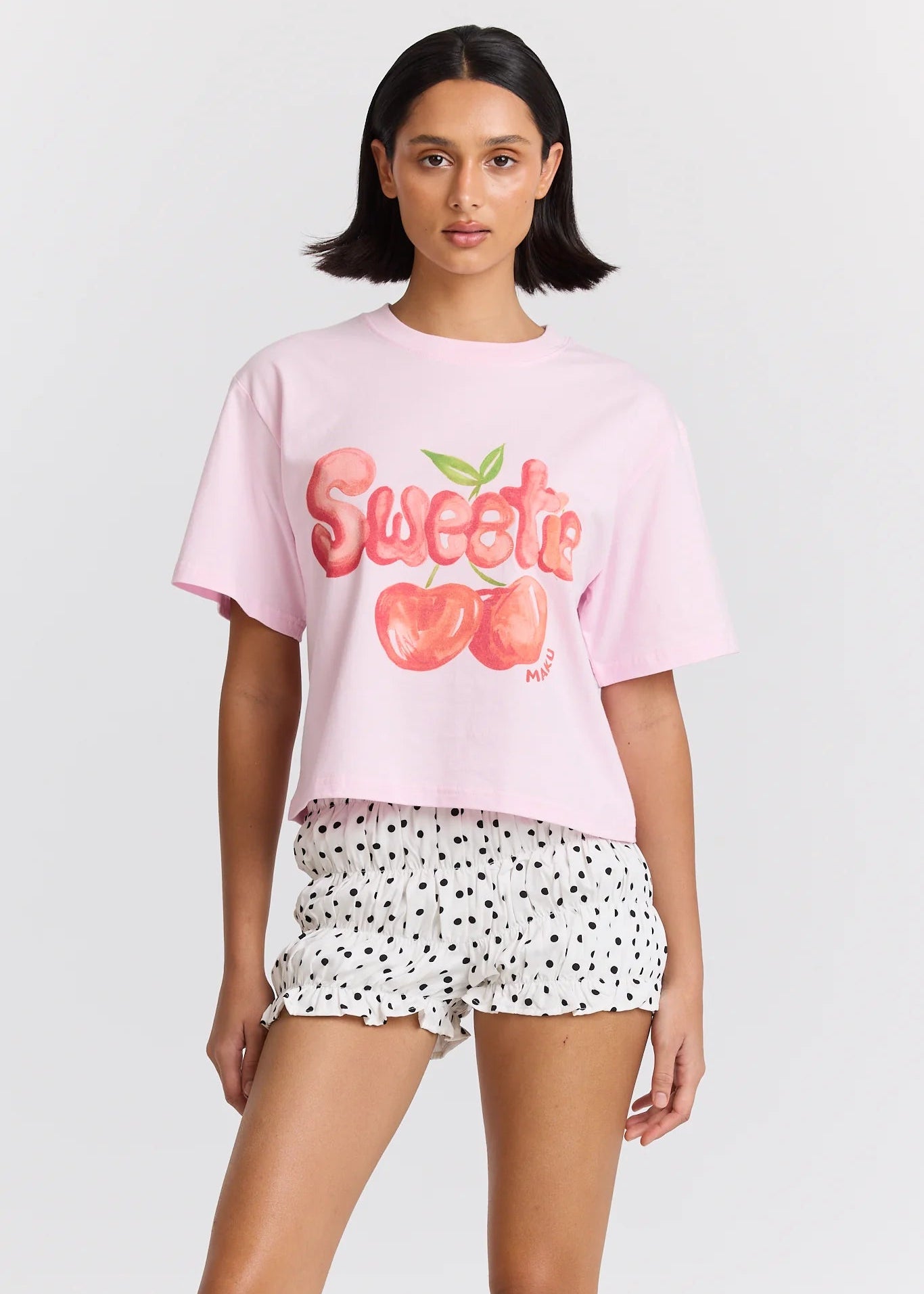 Woman wearing a pink 'Sweetie' t-shirt with cherry graphics and white shorts with black polka dots on a light gray background.