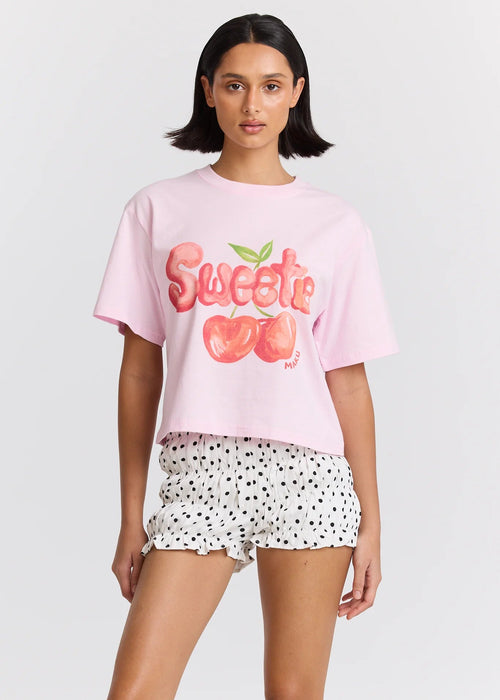 Woman wearing a pink 'Sweetie' t-shirt with cherry graphics and white shorts with black polka dots on a light gray background.