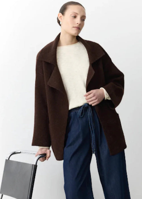 Woman wearing a brown coat and blue pants holding a chair against a white background