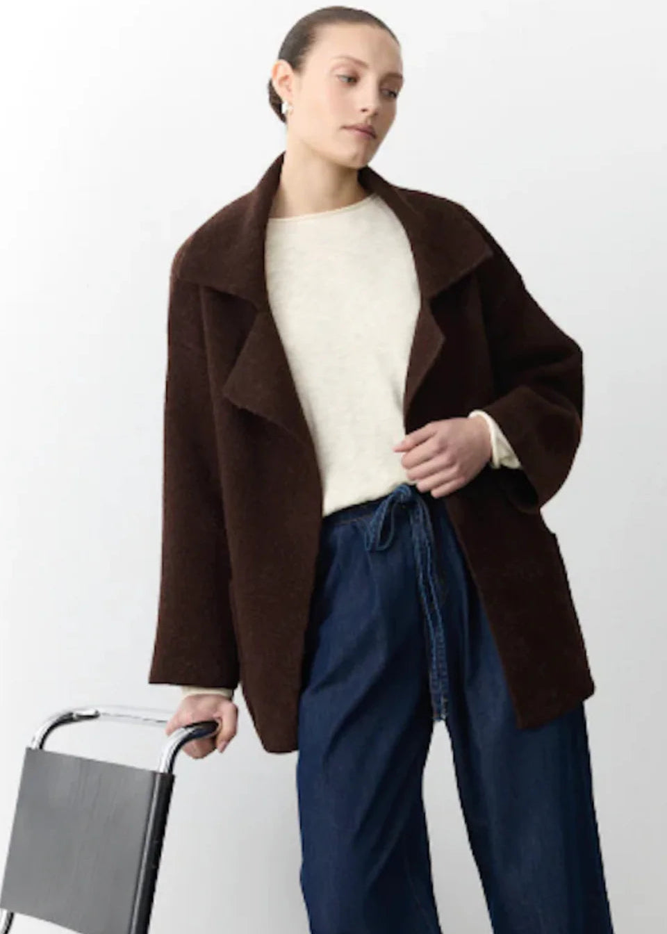 Woman wearing a brown coat and blue pants holding a chair against a white background