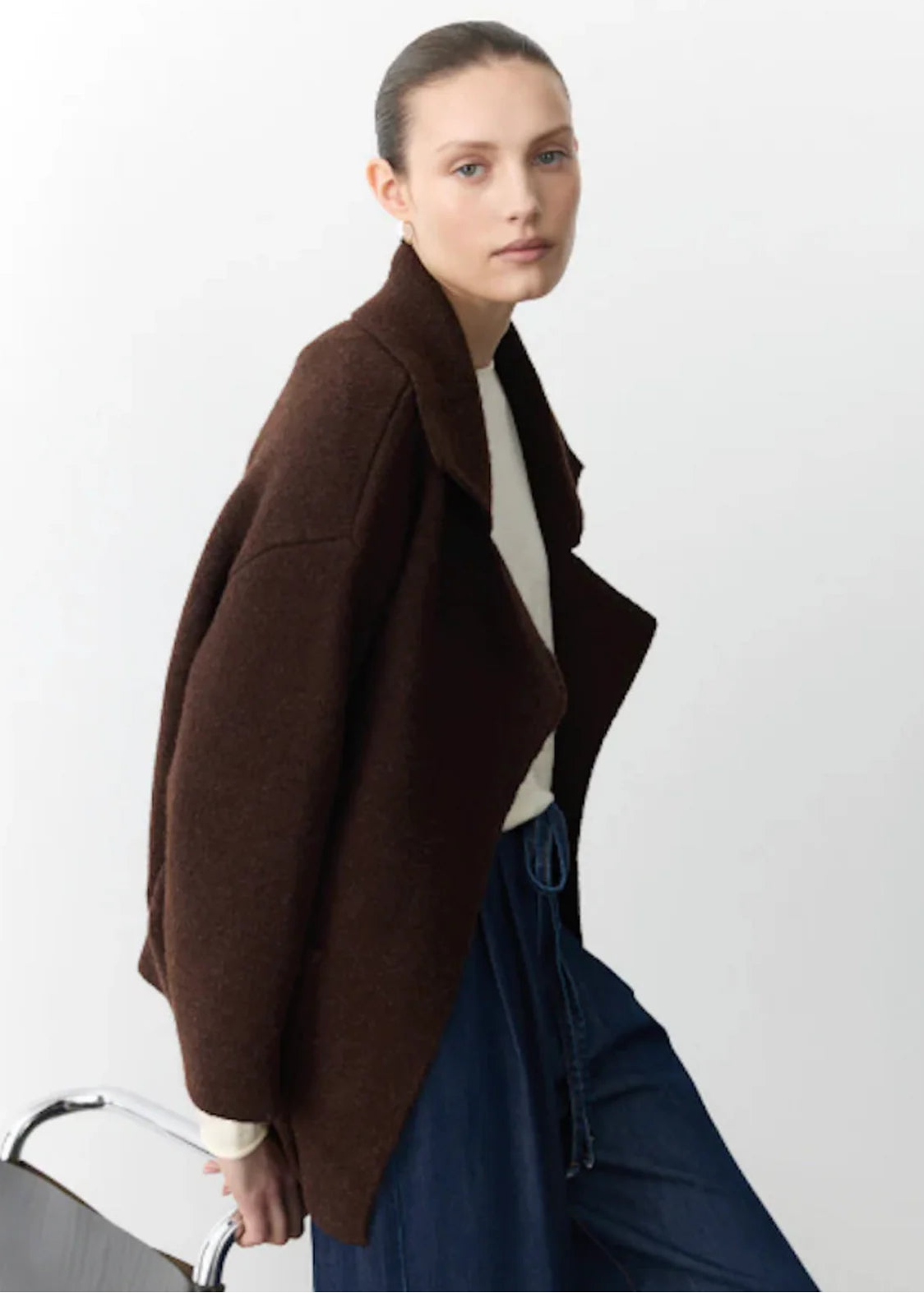 Woman wearing a brown coat and blue pants holding a chair against a white background