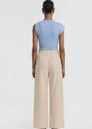 A model wearing a light blue knit top with a unique asymmetrical shoulder tie detail and beige drawstring trousers, shot against a plain light grey background.